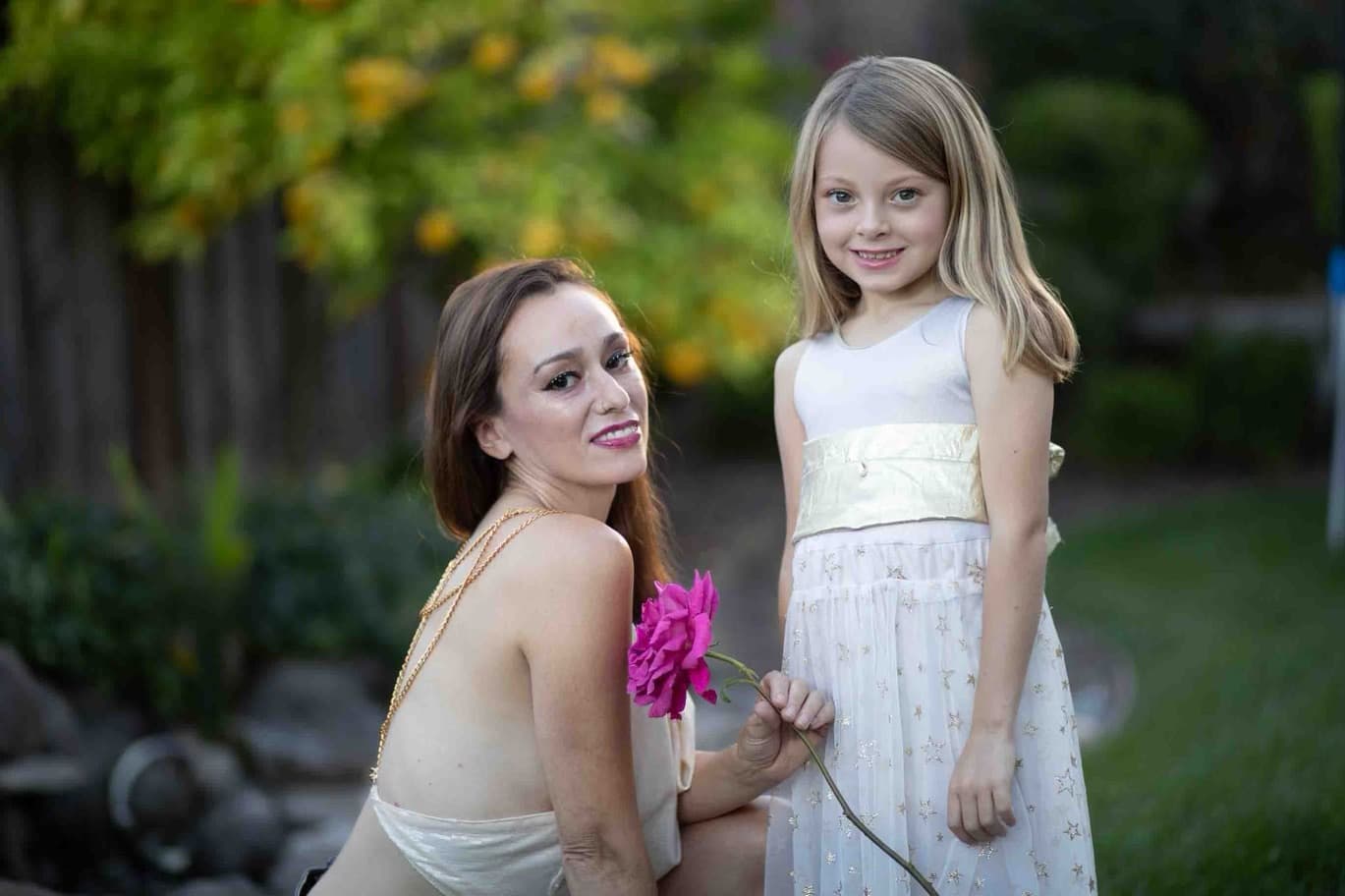 A woman in a cream dress kneels beside a young girl in a white party dress with yellow sash. The woman holds a pink rose. Garden setting with greenery.